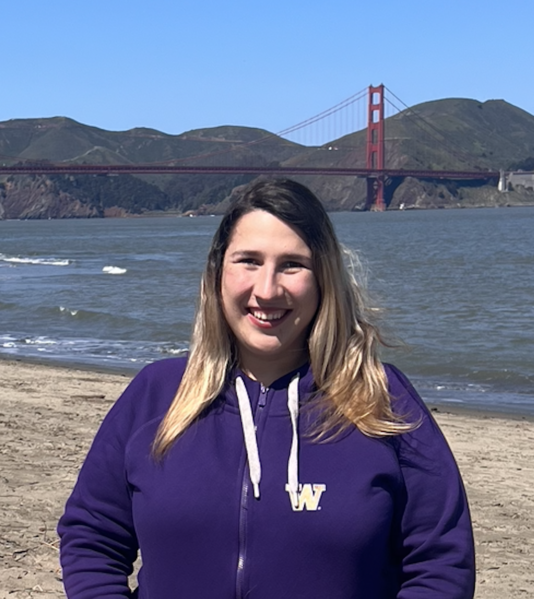 Kate stands in a purple UW sweater in front of Crissy Beach, a sandy California beach with the ocean glistening and the golden gate bridge in the background